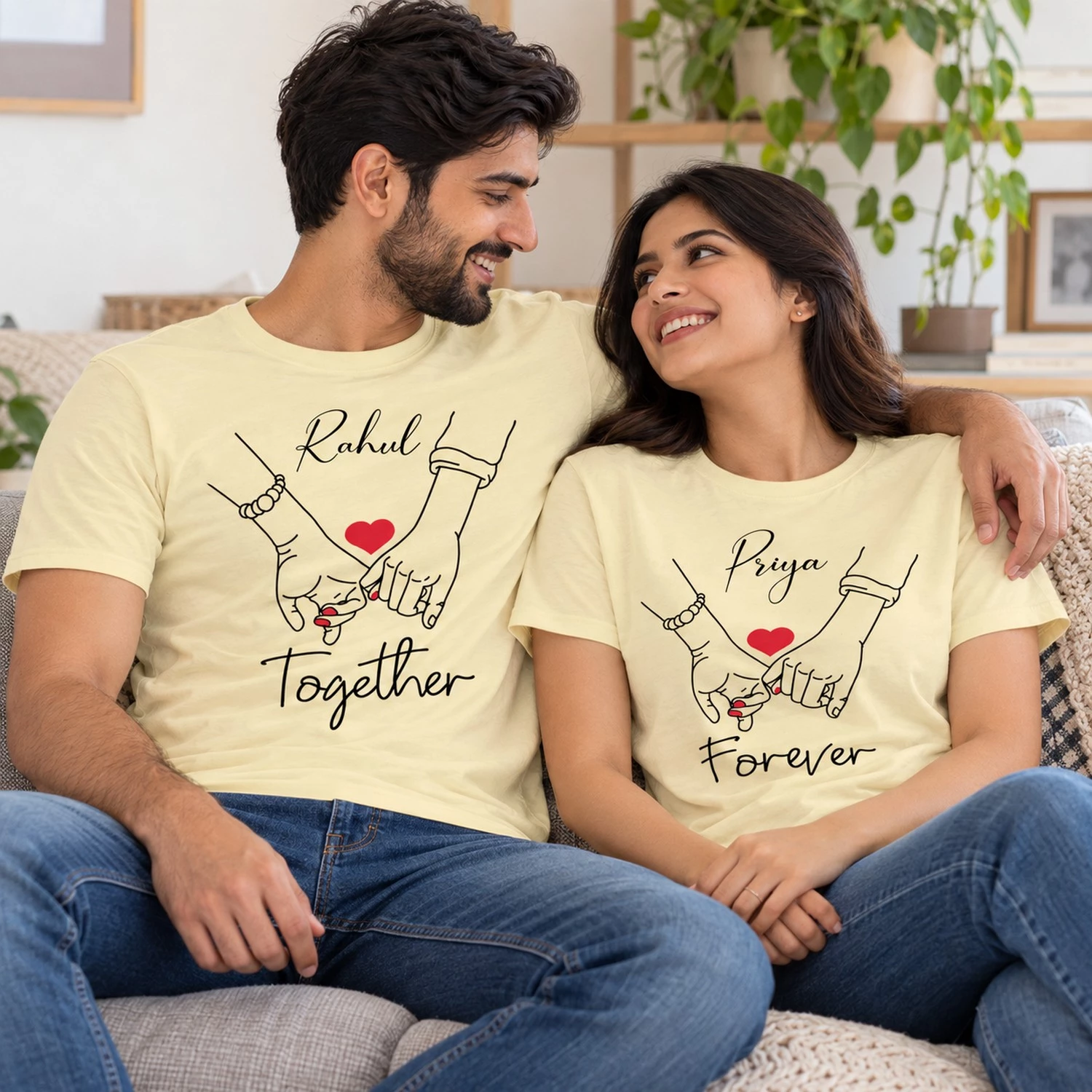 Smiling couple relaxing on a sofa in pale yellow personalized Rahul and Priya T-shirts with holding-hands artwork and Together Forever text.