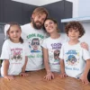 A happy family of four, including a mom, dad, big sister, and little brother, stands together in a kitchen wearing personalised family T-shirts. The white shirts feature cartoon avatars and customizable names, with the text "Cool Dad," "Cool Mom," "Big Sister," and "Little Brother" on them. This image highlights matching T-shirts for the whole family.