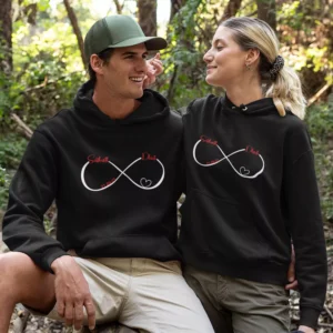 Matching black custom name split design couple hoodies with infinity heart print for couples sitting outdoors