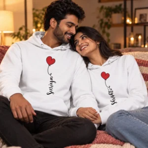 Couple sitting closely together in matching white personalized hoodies with red heart graphics and custom name prints in a warm indoor setting.