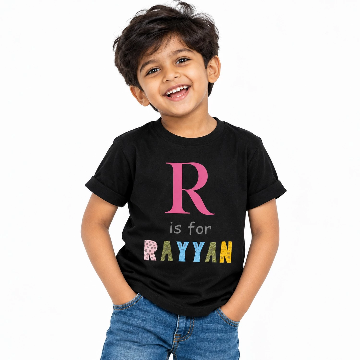 Smiling young boy wearing a black personalized T-shirt featuring a large pink letter R, the phrase is for, and the name RAYYAN in colorful patterned letters.