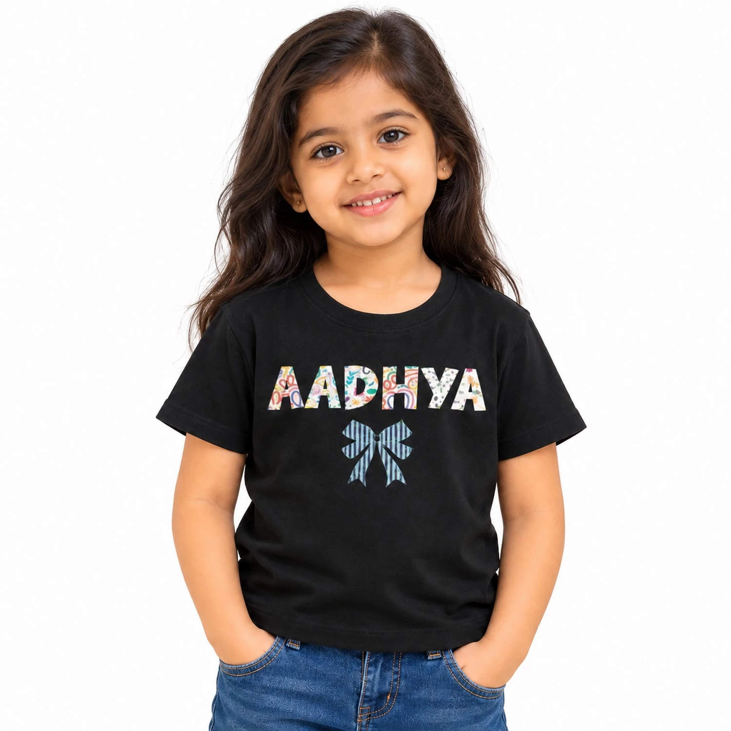 Young girl wearing a black personalized kids T-shirt featuring the name AADHYA in large multicolored patterned uppercase letters with a blue striped bow underneath.