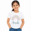 Smiling young girl wearing a white personalized kids T-shirt featuring a pastel rainbow with golden dots, pink and teal arches, a small heart and the name Aarohi in teal cursive lettering.