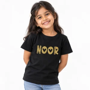 Smiling young girl wearing a black personalized T-shirt featuring the name NOOR in textured gold lettering with decorative heart-shaped cutouts.