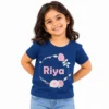Smiling young girl wearing a navy blue personalized T-shirt featuring the name Riya in pink letters surrounded by a circular wreath of pink roses, green leaves and delicate floral details.