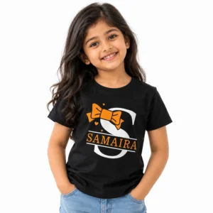 Smiling young girl wearing a black personalized T-shirt featuring a large white S monogram, orange bow, small hearts, and the name SAMAIRA in orange lettering.
