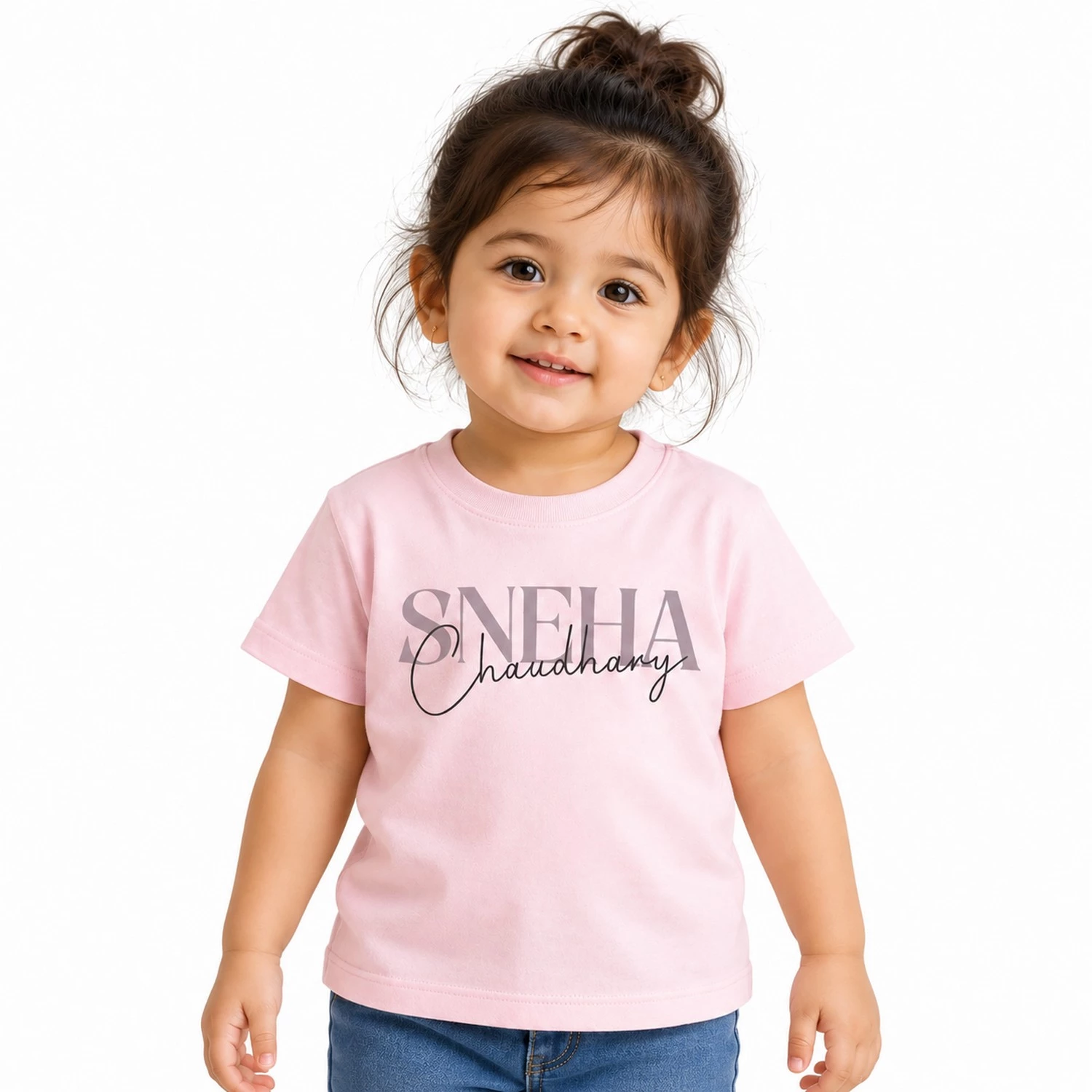 Young toddler girl wearing a light pink personalized T-shirt featuring the name SNEHA in large gray serif letters with Chaudhary overlaid in black cursive script.