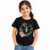 Young girl wearing a black personalized T-shirt featuring a large white T monogram, the name Trisha, and a circular floral wreath with pink flowers and green leaves.