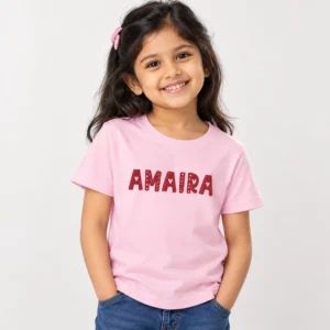 Light pink personalized girls' T-shirt with AMAIRA printed across the chest in dark red block letters decorated with small heart patterns.