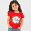 Red personalized kids T-shirt featuring the name Anika in black cursive text inside a white circle surrounded by colorful illustrated flowers.