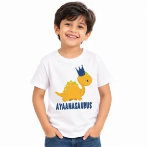 Smiling young boy wearing a white personalized kids T-shirt featuring a yellow cartoon dinosaur with a navy blue crown and the name AYAANASAURUS in bold blue lettering.
