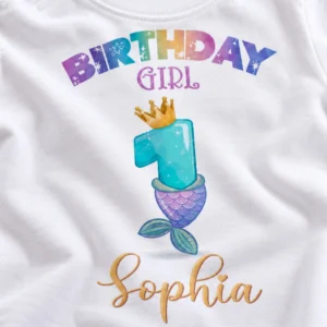 Close-up of white personalized first birthday T-shirt print showing colorful Birthday Girl lettering, turquoise number 1 with golden crown, purple mermaid tail, and Sophia in gold script.