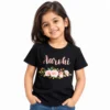 Smiling young girl wearing a black personalized kids T-shirt featuring the name Aarohi in soft pink cursive lettering above a floral arrangement of pink roses, white flowers, and green leaves.