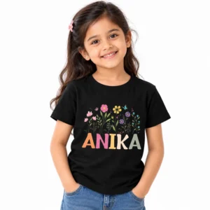 Smiling young girl wearing a black personalized kids T-shirt featuring colorful flowers, green stems, small butterflies, and the name ANIKA in large pastel multicolored block letters.