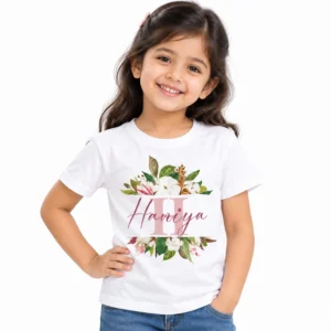 Smiling young girl wearing a white personalized kids T-shirt featuring a large pastel pink H monogram, watercolor flowers, green leaves, and the name Haniya in rose-colored cursive lettering.
