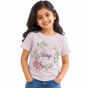 Smiling young girl wearing a pale blush pink personalized kids T-shirt featuring a large pastel pink N monogram, the name Navya in black script, and a wreath of green leaves and pink roses.