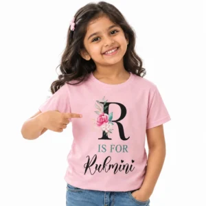 Smiling young girl wearing a light pink personalized T-shirt featuring a large black floral letter R, teal IS FOR lettering, and the name Rukmini in black cursive with small hearts.