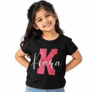 Smiling young Indian girl wearing a black personalized T-shirt with a large pink glitter-effect letter K and the name Kiara written across it in white cursive lettering.