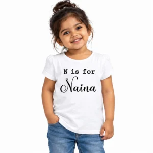 Smiling young Indian girl wearing a white personalized T-shirt with N is for printed in black typewriter-style lettering above the name Naina in decorative black cursive script.