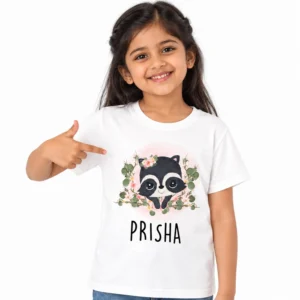Smiling Indian girl wearing a white personalized kids T-shirt with a cute black and white cartoon raccoon surrounded by pink flowers and green leaves, with PRISHA printed below.