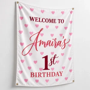 Personalized kids birthday tapestry banner with Amaira’s 1st birthday design, pink hearts, burgundy lettering, white fabric and four corner grommets.