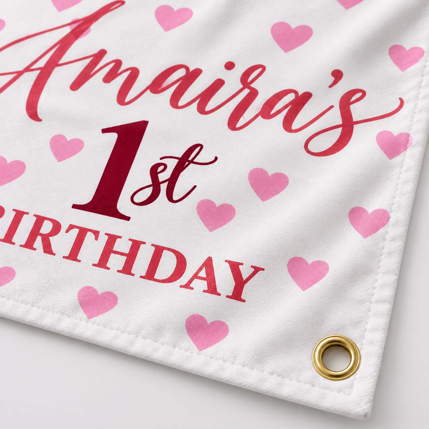 Close-up of a personalized kids birthday tapestry banner corner showing a gold metal grommet, white fabric texture, pink hearts and birthday print.