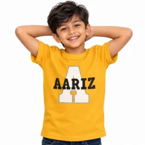 Smiling Indian boy wearing a yellow personalized kids T-shirt with a large white varsity-style letter A outlined in gray and the name AARIZ printed across it in bold black letters.