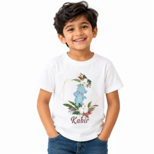 Smiling Indian boy wearing a white personalized T-shirt featuring a cute pastel blue teddy bear surrounded by floral leaves, white flowers and a gold geometric frame, with the name Kabir below.
