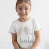 Child wearing white cotton T shirt with pastel rainbow design and custom name print