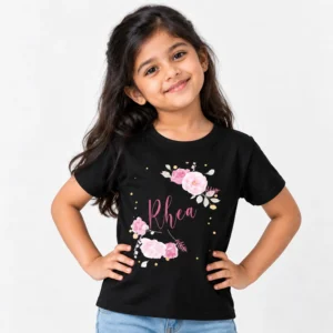 Personalized black kids T-shirt worn by a smiling girl, featuring the name Rhea in pink cursive lettering surrounded by pink roses and gold dots.