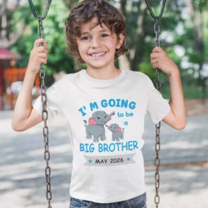 Smiling young boy wearing a big brother announcement T-shirt with cute elephant design and May 2026 reveal text while sitting on a swing