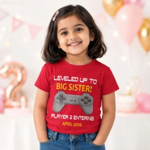 Little girl wearing a red big sister announcement t-shirt with “Player 2 Entering April 2026” gaming design