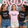 Three women wearing bride and team bride matching t-shirts at a bachelorette celebration with party decorations in the background
