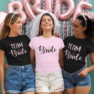 Three women wearing bride and team bride matching t-shirts at a bachelorette celebration with party decorations in the background
