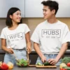 Couple wearing matching white t-shirts with Wife and Hubs EST design while cooking together in a kitchen