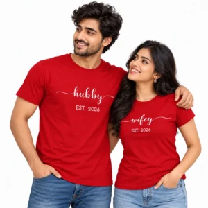 Smiling Indian couple wearing red matching T-shirts with white lowercase hubby and wifey cursive lettering, decorative flourishes, and EST. 2026 prints.