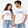Couple wearing matching white T-shirts with black WIFE and HUBS typography, outlined rectangular designs, small heart detail, and EST. 2026 prints.