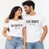 Smiling couple wearing matching white WIFEY and HUBBY T-shirts with bold black serif lettering and EST. 2026 printed beneath each design.