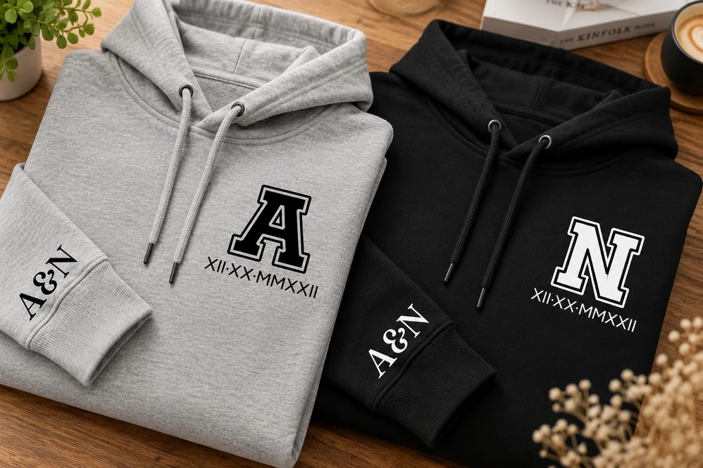 Folded grey and black couple hoodies featuring varsity-style A and N initials, Roman numeral dates and matching A&N sleeve prints.