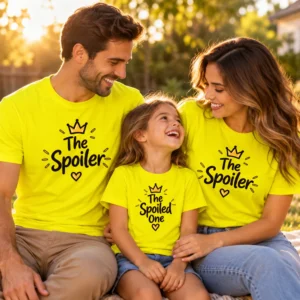 Parents and young daughter smiling together in warm golden-hour sunlight while wearing matching yellow family T-shirts featuring The Spoiler and The Spoiled One crown and heart designs.