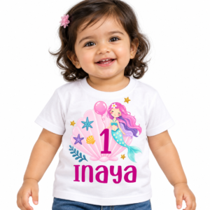Indian toddler wearing a white personalized 1st birthday kids t-shirt with a pink mermaid shell design, number 1, and the name Inaya on a plain light background
