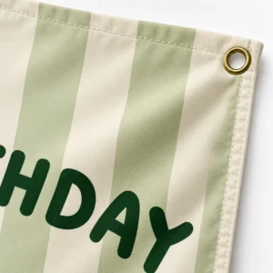 Close-up of a personalized birthday tapestry corner showing a gold-tone metal grommet, stitched fabric edge, sage stripes, and dark green printed lettering.