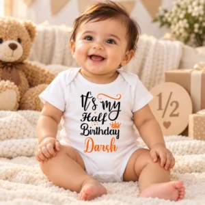 Smiling baby in a white personalized Darsh half birthday romper surrounded by soft birthday props, gifts and a wooden half milestone sign.