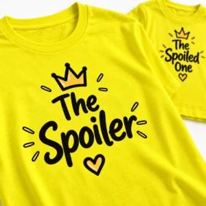 Close-up of a bright yellow The Spoiler family T-shirt featuring bold black handwritten lettering, a crown illustration, decorative accents, and a heart, with a matching children's T-shirt in the background.